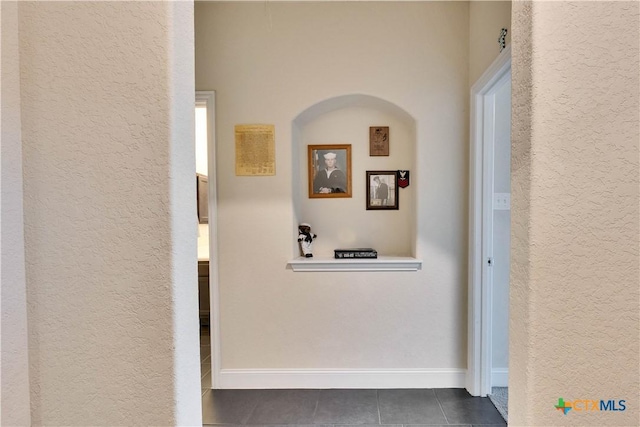 hall featuring dark tile patterned flooring, a textured wall, and baseboards