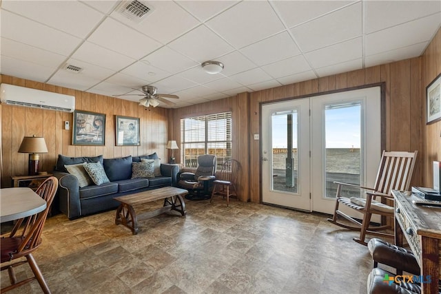 living room with a water view, a paneled ceiling, an AC wall unit, wooden walls, and ceiling fan