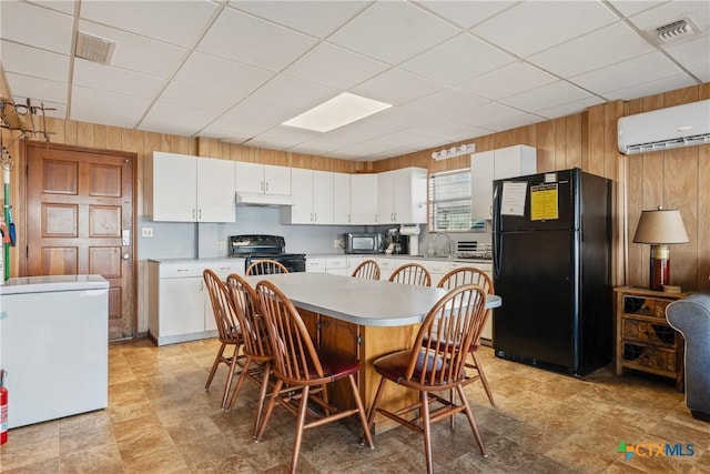 kitchen with black appliances, a wall mounted AC, a drop ceiling, wooden walls, and white cabinets