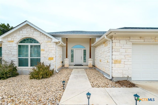 view of exterior entry with a garage, stone siding, and a shingled roof