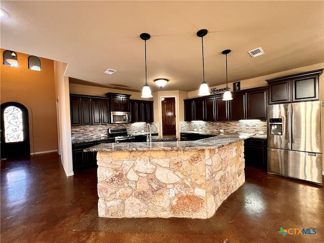 kitchen featuring stainless steel appliances, decorative backsplash, dark brown cabinetry, an island with sink, and decorative light fixtures