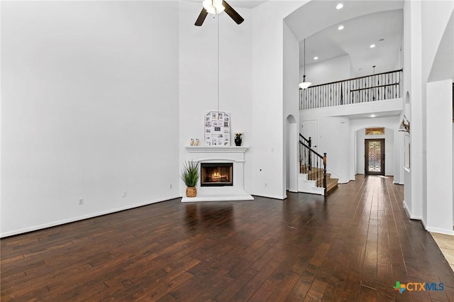 unfurnished living room featuring hardwood / wood-style flooring, a warm lit fireplace, stairway, arched walkways, and ceiling fan