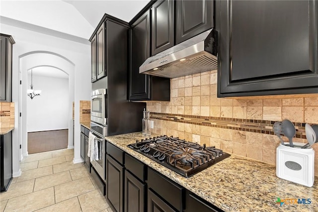 kitchen featuring dark cabinetry, light stone countertops, arched walkways, appliances with stainless steel finishes, and under cabinet range hood