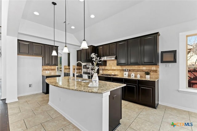 kitchen with a kitchen island with sink, a sink, light stone counters, under cabinet range hood, and backsplash