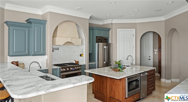 kitchen featuring high quality appliances, premium range hood, a kitchen island with sink, sink, and light stone countertops