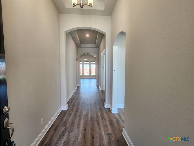 hallway with baseboards, arched walkways, dark wood-type flooring, an inviting chandelier, and a tray ceiling