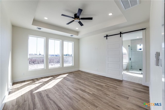 unfurnished bedroom featuring a raised ceiling, ensuite bath, ceiling fan, a barn door, and light hardwood / wood-style floors