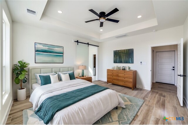 bedroom with a raised ceiling, a barn door, ceiling fan, and light wood-type flooring