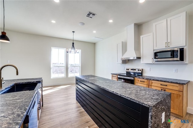 kitchen featuring wall chimney exhaust hood, dark stone counters, stainless steel appliances, decorative light fixtures, and white cabinetry