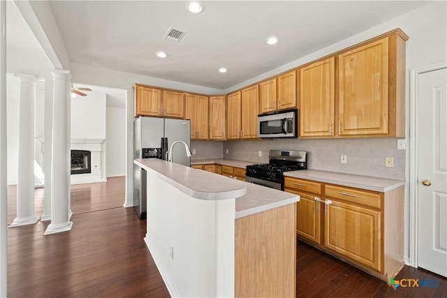 kitchen with dark wood-type flooring, a center island with sink, decorative columns, ceiling fan, and appliances with stainless steel finishes