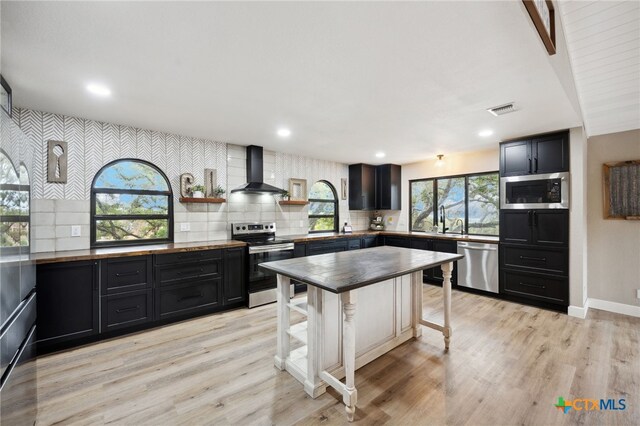 30817 Wildcat Dr, Bulverde, TX 78163 - kitchen featuring stainless steel appliances, wall chimney range hood, dark cabinets, backsplash, and light wood-style flooring