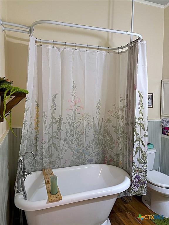 full bath with a wainscoted wall, toilet, ornamental molding, wood finished floors, and a soaking tub