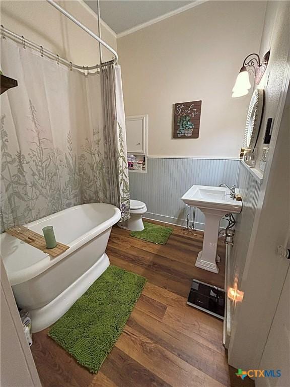 bathroom with wood finished floors, a soaking tub, wainscoting, crown molding, and toilet