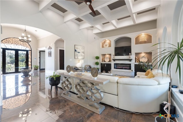 living room with arched walkways, a towering ceiling, a glass covered fireplace, a chandelier, and coffered ceiling