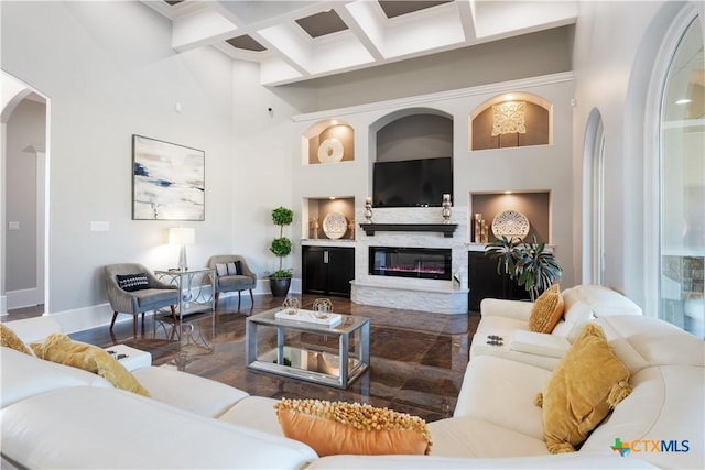 living room featuring arched walkways, coffered ceiling, a towering ceiling, baseboards, and a glass covered fireplace