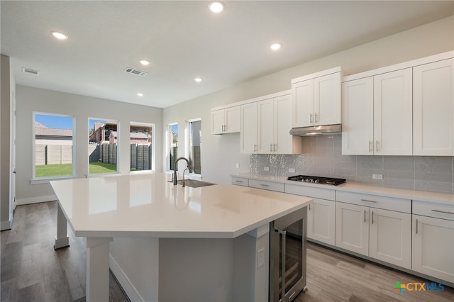 kitchen with light wood-type flooring, white cabinets, stainless steel gas stovetop, beverage cooler, and a kitchen island with sink