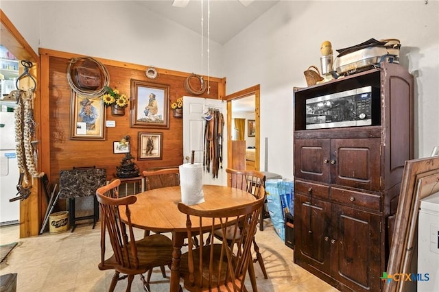 dining space with light floors, ceiling fan, wood walls, and high vaulted ceiling
