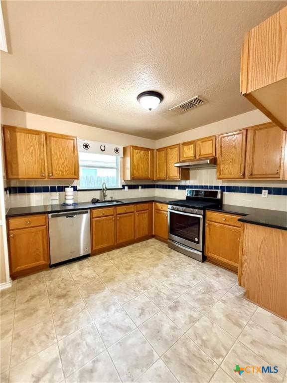 1828 Winchester Dr, Temple, TX 76502 - kitchen featuring appliances with stainless steel finishes, dark countertops, brown cabinetry, a textured ceiling, and under cabinet range hood