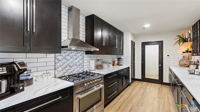 kitchen featuring high end range, decorative backsplash, wall chimney range hood, light stone countertops, and light hardwood / wood-style flooring