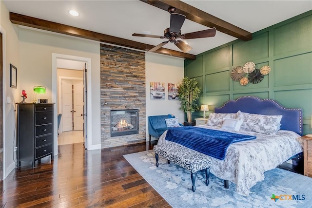 bedroom with beamed ceiling, ceiling fan, a stone fireplace, and dark wood-type flooring