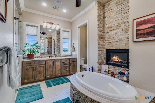 bathroom with a stone fireplace, ornamental molding, vanity, a relaxing tiled tub, and ceiling fan