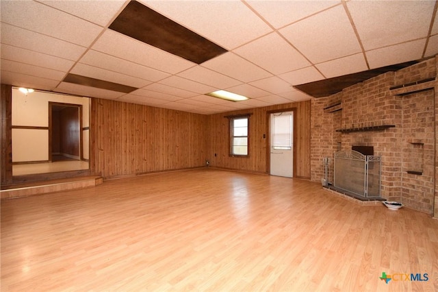 unfurnished living room featuring a paneled ceiling, light wood-style floors, a brick fireplace, and wooden walls