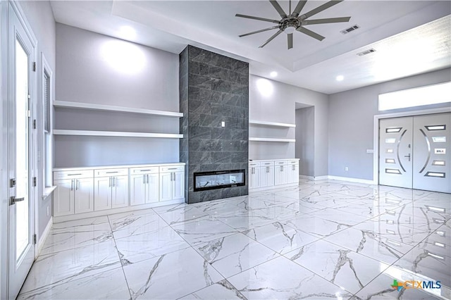 unfurnished living room with a fireplace, built in shelves, a tray ceiling, and ceiling fan