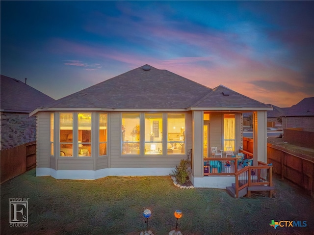 back of property at dusk featuring a shingled roof, fence, and a lawn