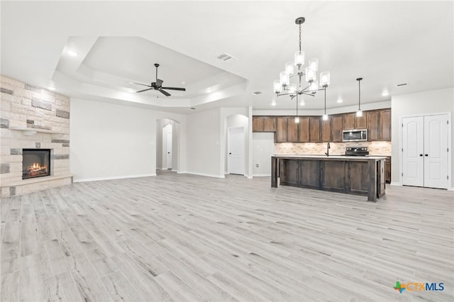 unfurnished living room with light hardwood / wood-style flooring, a stone fireplace, ceiling fan with notable chandelier, and a raised ceiling