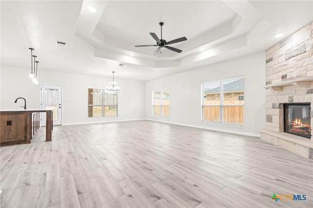 unfurnished living room featuring light wood-type flooring, ceiling fan with notable chandelier, a fireplace, and a tray ceiling