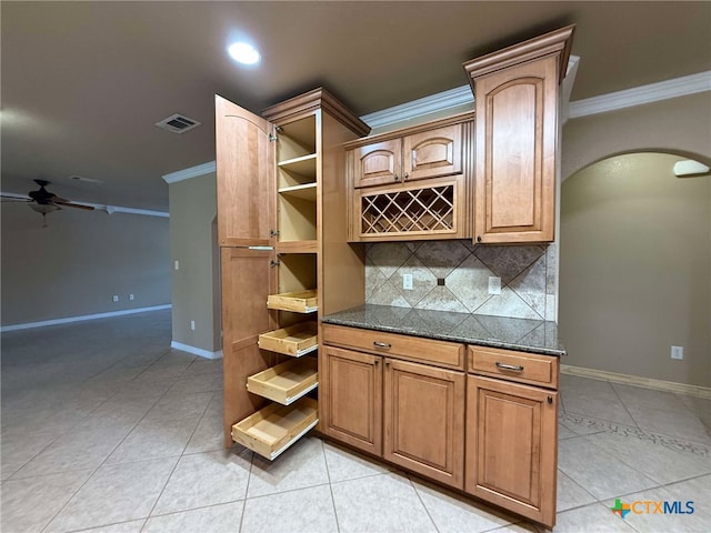 kitchen with light tile patterned flooring, crown molding, dark stone countertops, ceiling fan, and backsplash