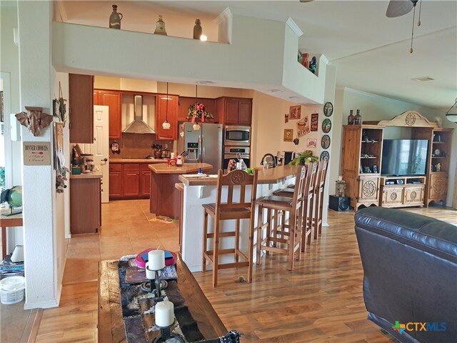 kitchen with appliances with stainless steel finishes, light wood-type flooring, wall chimney exhaust hood, a breakfast bar, and ceiling fan