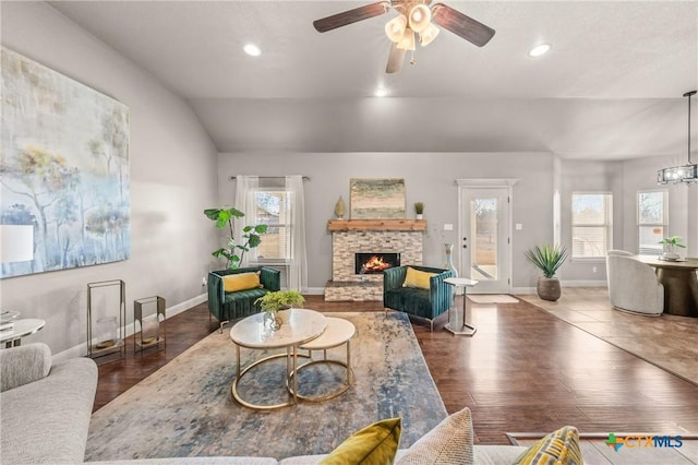 living room with dark wood-type flooring, lofted ceiling, a stone fireplace, and ceiling fan with notable chandelier