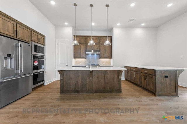 kitchen featuring a kitchen island with sink, wood finished floors, light countertops, under cabinet range hood, and appliances with stainless steel finishes