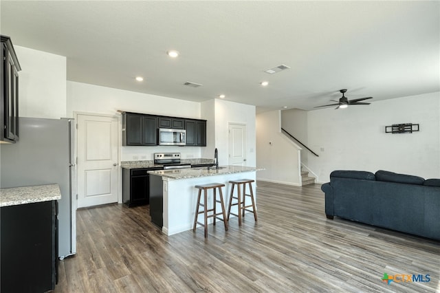 kitchen featuring open floor plan, a center island with sink, appliances with stainless steel finishes, wood finished floors, and a sink