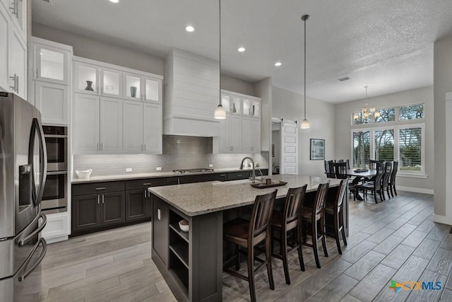 kitchen featuring appliances with stainless steel finishes, glass insert cabinets, a center island with sink, and white cabinets