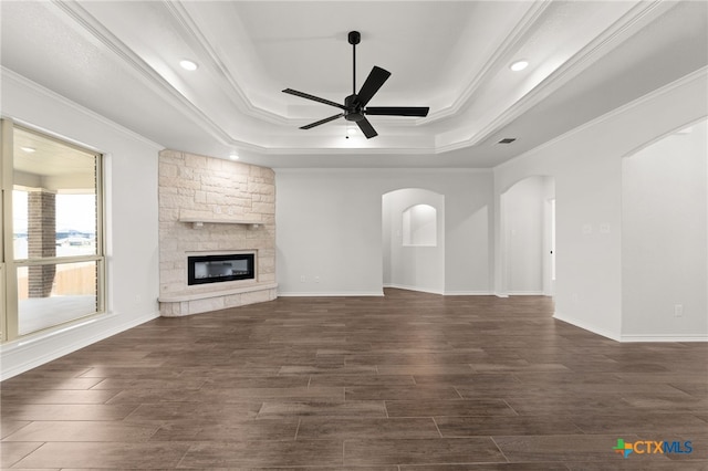 unfurnished living room with ornamental molding, ceiling fan, a tray ceiling, a stone fireplace, and dark wood-type flooring