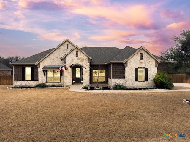 french country style house with a shingled roof, fence, a front lawn, and brick siding