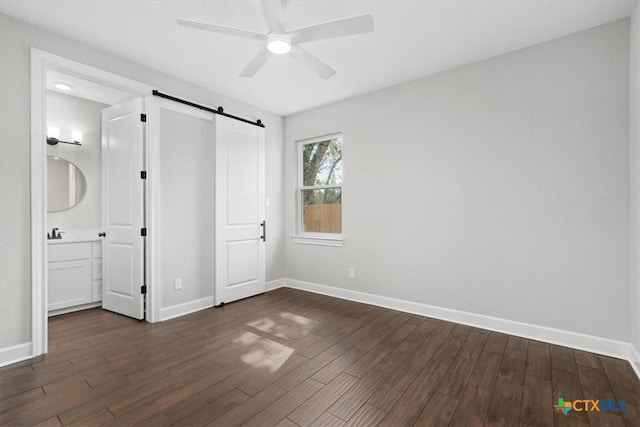 unfurnished bedroom featuring ensuite bathroom, ceiling fan, dark wood-type flooring, and a barn door