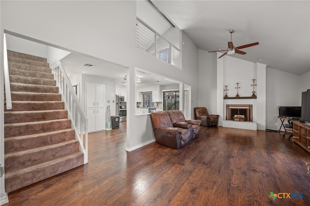 living room featuring ceiling fan, a brick fireplace, a towering ceiling, and dark hardwood / wood-style flooring