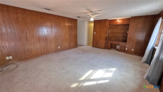 unfurnished living room featuring wood walls, light carpet, ceiling fan, built in desk, and a textured ceiling