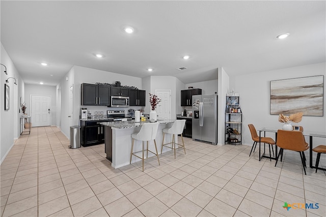 kitchen with a kitchen bar, stainless steel appliances, light stone counters, light tile patterned floors, and an island with sink