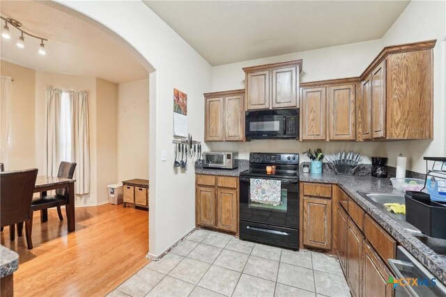 kitchen with brown cabinetry, light tile patterned flooring, arched walkways, black appliances, and dark countertops