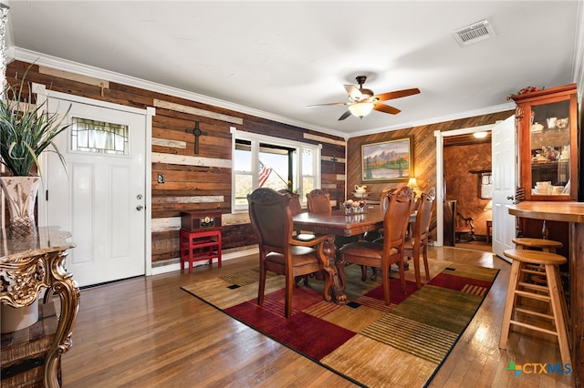 dining space with wood walls, ceiling fan, crown molding, and dark wood-type flooring