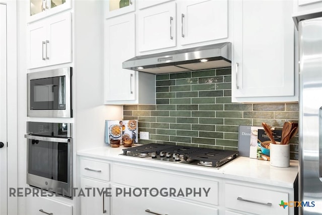 kitchen with appliances with stainless steel finishes, under cabinet range hood, white cabinetry, tasteful backsplash, and glass insert cabinets
