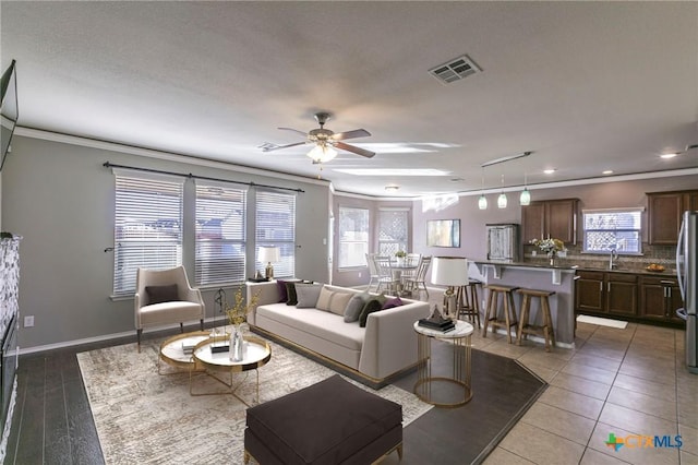 living area with light tile patterned floors, baseboards, visible vents, ceiling fan, and crown molding