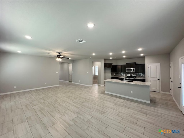 kitchen featuring sink, light stone counters, a center island with sink, ceiling fan, and backsplash