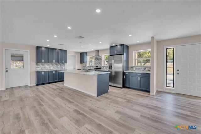 kitchen featuring wall chimney exhaust hood, a kitchen island with sink, light wood-type flooring, stainless steel refrigerator with ice dispenser, and backsplash