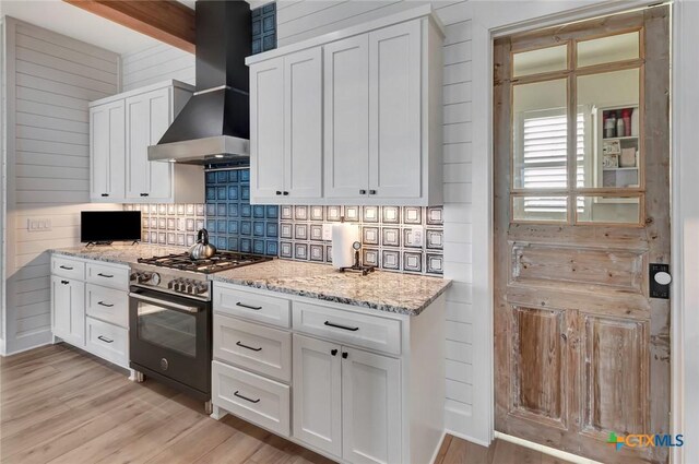 kitchen with wall chimney range hood, high end range, light stone counters, light hardwood / wood-style flooring, and white cabinets