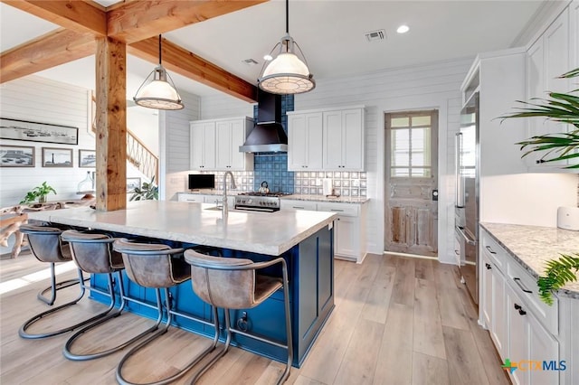 kitchen with pendant lighting, a kitchen island with sink, wall chimney exhaust hood, light wood-type flooring, and white cabinetry